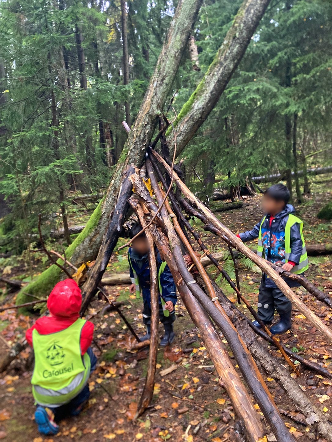 Olari children exploring a woodland den made from branches, with visible faces gently blurred